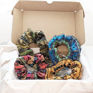 May include: Four colorful patterned hair scrunchies in a cardboard box. The scrunchies are made of fabric and have a variety of colors and patterns, including blue, green, yellow, and red.