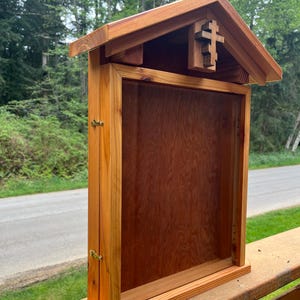 May include: A wooden display case with a pitched roof and a cross-shaped cutout. The case is made of light brown wood and has a rectangular opening. The case is designed to be mounted on a wall or placed on a flat surface.