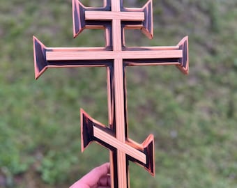 May include: A wooden cross with a dark brown stain and a light wood grain. The cross is made of multiple pieces of wood that are joined together. The cross is a traditional Orthodox Christian cross.