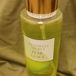 May include: A clear glass bottle of Victoria's Secret Pear Glace body spray with a gold cap. The bottle is filled with a yellow liquid and has a white label with the text "Victoria's Secret Pear Glace" printed on it.