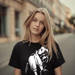 Op de afbeelding: Zwart T-shirt met een witte afbeelding van een persoon in een zittende pose, met een bedachtzame uitdrukking. De persoon op de afbeelding draagt een zwart T-shirt met een witte afbeelding. De achtergrond is een vage straatscène.