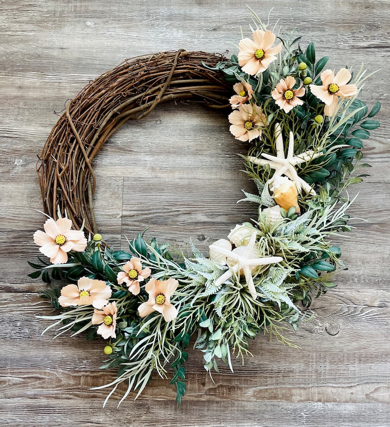 May include: A grapevine wreath decorated with peach-colored flowers and white seashells. The wreath is surrounded by green leaves and vines.