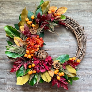 May include: A fall-themed wreath with a grapevine base, adorned with green magnolia leaves, orange hydrangeas, burgundy leaves, orange berries, and small pinecones. The wreath has a rustic, natural aesthetic, perfect for seasonal home decor.