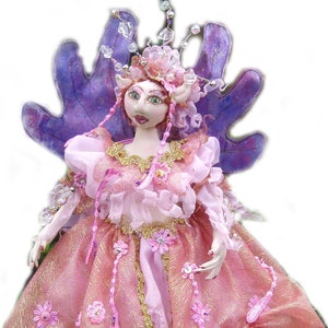 Puede incluir: Muñeca de hada artesanal con un vestido rosa y dorado, adornada con detalles florales y abalorios. La muñeca presenta alas moradas y una corona enjoyada. La muñeca está sobre una esfera morada.