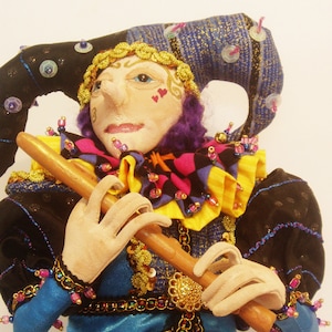 Puede incluir: Una muñeca hecha a mano vestida de bufón, con un traje azul, dorado y negro, una peluca morada y un gran gorro de bufón azul y dorado. La muñeca sostiene una flauta de madera y tiene la cara pintada con corazones rosas.