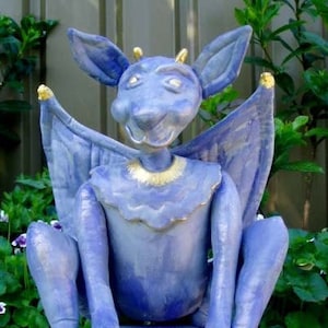 May include: A blue gargoyle statue with gold detailing. The statue features large ears, wings, and holds a purple orb. The gargoyle is seated on a blue base, with gold accents on the claws and collar.