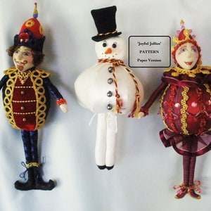 Puede incluir: Tres muñecas de tela caprichosas. La primera muñeca viste un atuendo azul y rojo con adornos dorados y un sombrero alto. La segunda muñeca es un muñeco de nieve con un sombrero de copa negro. La tercera muñeca está vestida con un atuendo rojo y dorado con una corona. El texto de la imagen dice 'Joyful Jollies' PATTERN Paper Version.