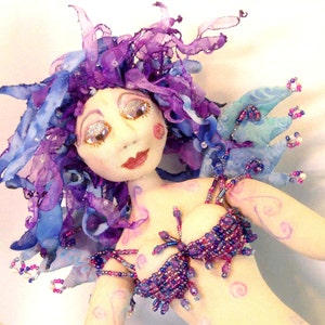 Może przedstawiać: Ręcznie robiona lalka z fioletowymi i niebieskimi włosami, korpusem ozdobionym koralikami i figlarnym wyrazem twarzy.