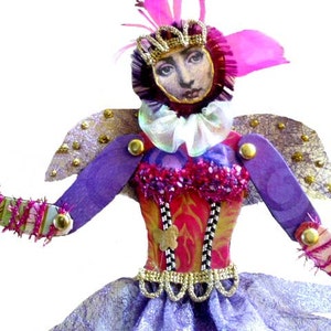 May include: A purple and gold fairy doll with pink and white wings, a pink crown, and a pink and white ruffled collar. The doll is wearing a pink and gold dress with a black and white pattern and a gold belt.