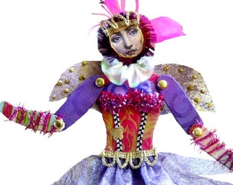 May include: A purple and gold fairy doll with pink and white wings, a pink crown, and a pink and white ruffled collar. The doll is wearing a pink and gold dress with a black and white pattern and a gold belt.