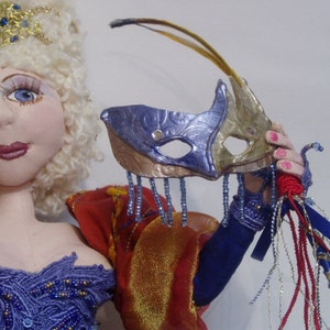 May include: A doll wearing a white wig, a blue and gold mask, a red and gold dress, and blue and gold beaded jewelry. The doll is holding a red and gold whip.