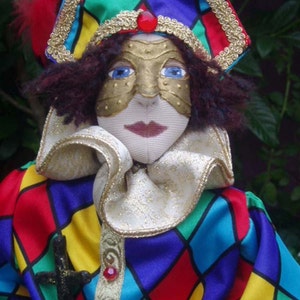 May include: A colorful jester doll with a red, green, yellow, and blue diamond pattern on its costume. The doll is wearing a gold mask with a red jewel on its forehead and a gold and red hat with a feather.