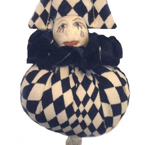 May include: A black and white checkered fabric jester doll with a white tassel. The doll has a painted face with tear drops and a red mouth.