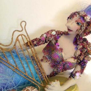 Op de afbeelding: Handgemaakte zeemeerminfiguur met een gouden harp. De zeemeermin heeft een blauw-paarse gevederde staart, een kralen lijfje en een hoofddeksel met schelpen en veren. Een uniek kunstwerk.