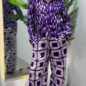 May include: A two-piece purple and white patterned outfit. The long-sleeved button-down shirt features a white feather-like design. The wide-leg pants have a white square pattern. The outfit is worn by a person with gold earrings and a bun.