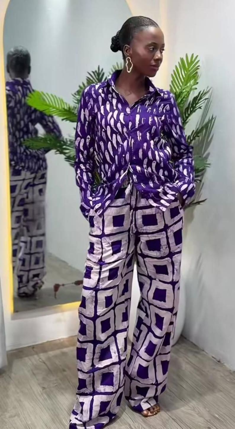 May include: A two-piece purple and white patterned outfit. The long-sleeved button-down shirt features a white feather-like design. The wide-leg pants have a white square pattern. The outfit is worn by a person with gold earrings and a bun.