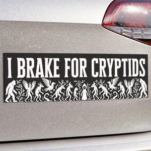 Peut inclure: Autocollant de pare-chocs rectangulaire noir avec le texte blanc "I BRAKE FOR CRYPTIDS". Une illustration blanche de divers cryptides, dont des créatures ailées et des figures humanoïdes, est en dessous. L'autocollant est sur une voiture argentée.