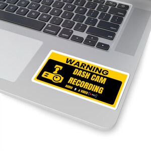May include: A rectangular sticker with a yellow and black design, featuring the text "WARNING DASH CAM RECORDING" and a camera icon. The sticker also includes "AUDIO & VIDEO [REC]" text. The sticker is placed on a laptop.
