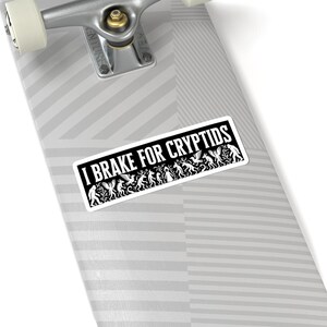 May include: A white sticker with the text "I Brake for Cryptids" in a black rectangular frame. The sticker features a line of stylized cryptid illustrations in black and white, including winged creatures and other mythical beings.