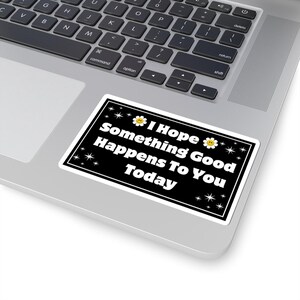 Pu&ograve; includere: Un adesivo rettangolare nero con bordo bianco. Presenta il testo "I Hope Something Good Happens To You Today" in bianco, con grafica di margherite e stelle. L'adesivo &egrave; posizionato su un computer portatile.