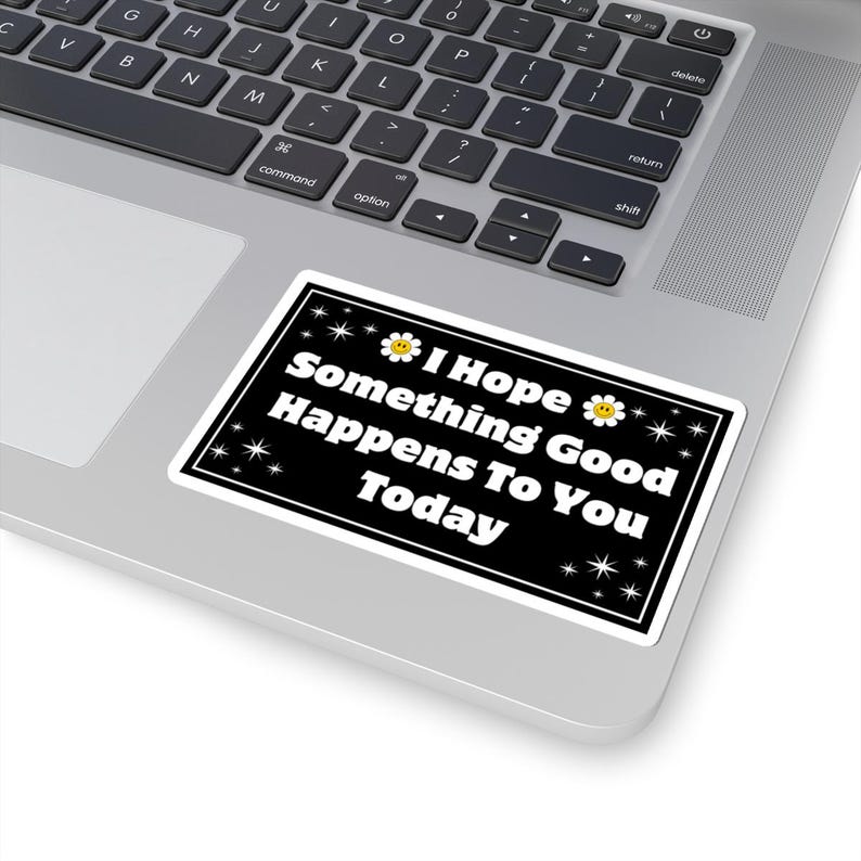 Pu&ograve; includere: Un adesivo rettangolare nero con bordo bianco. Presenta il testo "I Hope Something Good Happens To You Today" in bianco, con grafica di margherite e stelle. L'adesivo &egrave; posizionato su un computer portatile.