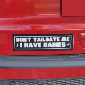 May include: A rectangular bumper sticker with the text "DON'T TAILGATE ME I HAVE RABIES" in white letters on a black background. The sticker is affixed to a red vehicle.