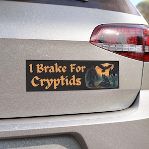 Peut inclure: Autocollant de pare-chocs rectangulaire noir avec le texte "I Brake For Cryptids" en lettres orange. L'autocollant présente une illustration de Bigfoot, d'un loup et d'un oiseau. L'autocollant est apposé à l'arrière d'une voiture argentée.