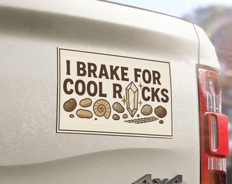 May include: A rectangular bumper sticker with a cream background and a dark brown border. The text reads I BRAKE FOR COOL ROCKS in dark brown letters. Below the text are illustrations of various brown rocks, a fossil, and a crystal.