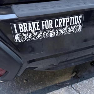 May include: A white bumper sticker on a black car bumper. The sticker reads "I BRAKE FOR CRYPTIDS" in bold white letters. Below the text is a white silhouette design of various cryptids.