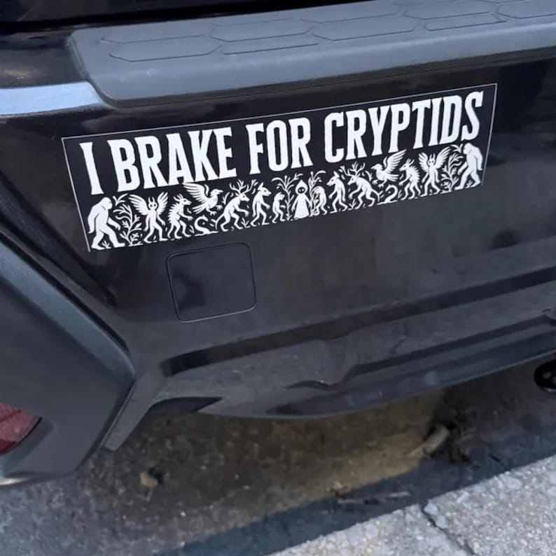 May include: A white bumper sticker on a black car bumper. The sticker reads "I BRAKE FOR CRYPTIDS" in bold white letters. Below the text is a white silhouette design of various cryptids.