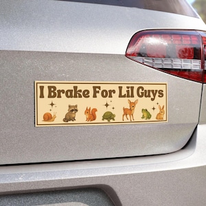 May include: A rectangular bumper sticker with a cream background and brown text that reads I Brake For Lil Guys. The design features small illustrations of a snail, raccoon, squirrel, turtle, deer, frog, and rabbit, accented by four-pointed stars.