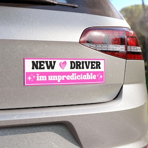 May include: A rectangular bumper sticker with the text "NEW ♥ DRIVER im unpredictable" in black and pink. The sticker is affixed to the back of a silver car, with the taillight visible.