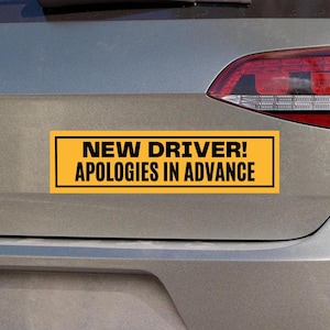 Op de afbeelding: Een gele en zwarte autobumper sticker met de tekst "NEW DRIVER! APOLOGIES IN ADVANCE".