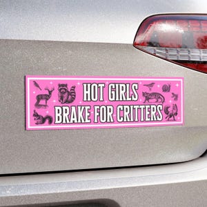 Peut inclure: Autocollant de pare-chocs rose avec le texte "HOT GIRLS BRAKE FOR CRITTERS" en blanc. L'autocollant présente des illustrations d'animaux, dont un raton laveur, un cerf, un écureuil et un renard. L'autocollant est fixé à l'arrière d'une voiture argentée.