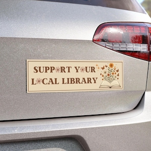 Puede incluir: Una pegatina rectangular para parachoques con el texto "SUPPORT YOUR LOCAL LIBRARY" en letras marrones. La pegatina presenta ilustraciones florales y de mariposas. La pegatina está adherida a la parte trasera de un coche plateado.