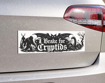 May include: Black and white bumper sticker with the text "I Brake for Cryptids." The sticker features illustrations of a Bigfoot, a Loch Ness Monster, and other cryptids. The sticker is rectangular and mounted on a silver car.