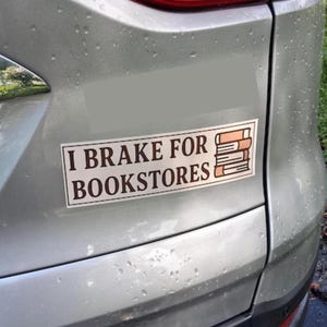 Puede incluir: Una pegatina rectangular en un coche plateado con el texto "I BRAKE FOR BOOKSTORES" en letras marrones. Una pila de libros está a la derecha del texto. La pegatina tiene un fondo blanco.