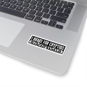 May include: A rectangular sticker with the text "I BRAKE FOR CRYPTIDS" in white on a black background. The sticker features various cryptid silhouettes. The sticker is placed on a silver laptop.