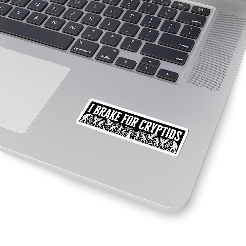 May include: A rectangular sticker with the text "I BRAKE FOR CRYPTIDS" in white on a black background. The sticker features various cryptid silhouettes. The sticker is placed on a silver laptop.