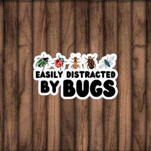 Peut inclure: Un autocollant blanc avec le texte "EASILY DISTRACTED BY BUGS" et des illustrations d'insectes. Les insectes comprennent un coléoptère vert, une coccinelle rouge, une abeille, un coléoptère brun et une mouche bleue. Le fond est en bois.