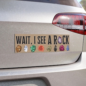 May include: A rectangular bumper sticker with the text "WAIT, I SEE A ROCK" in black. Below the text are illustrations of various colorful gemstones, including brown, clear, green, red, gold, black, purple, and blue.