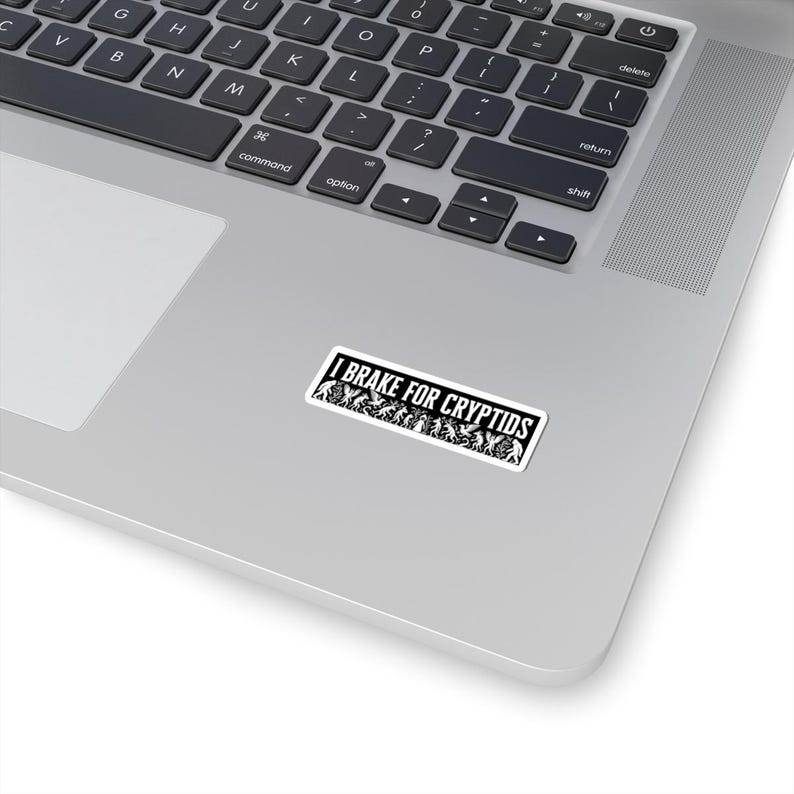 May include: A rectangular white sticker with black text that reads "I BRAKE FOR CRYPTIDS." The sticker features a silhouette of various cryptids. The sticker is placed on a silver laptop.