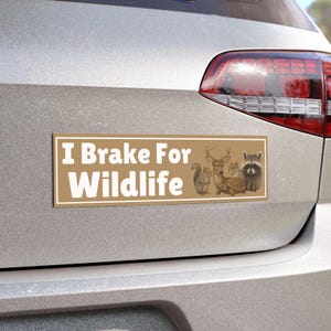 May include: A rectangular bumper sticker with a tan background and a white border. The text reads I Brake For Wildlife in bold white letters. To the right, there are illustrations of a squirrel, a deer, a chipmunk, a turtle, and a raccoon.