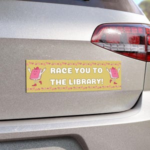 Peut inclure: Autocollant de pare-chocs jaune avec le texte "RACE YOU TO THE LIBRARY!" et des personnages de livres de dessins animés. L'autocollant est rectangulaire et possède une bordure décorative. Il est fixé à l'arrière d'une voiture argentée.