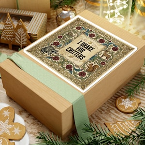 May include: A wooden gift box with a sticker that reads "I Brake for Critters." The sticker features a raccoon, squirrel, and deer, surrounded by floral designs. A light green ribbon wraps around the box, and gingerbread cookies and pine needles are visible.