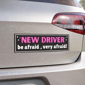 May include: A black rectangular bumper sticker with pink text reading "NEW DRIVER" and white text saying "be afraid, very afraid!". The sticker is affixed to a silver car.