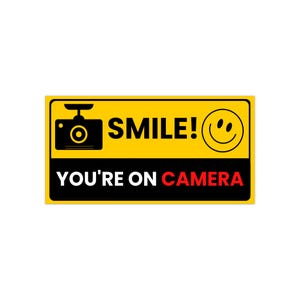 Puede incluir: Un letrero rectangular amarillo y negro con el texto "SMILE!" y "YOU'RE ON CAMERA". El letrero también presenta un icono de cámara y una cara sonriente.
