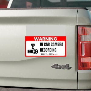 Può includere: Un adesivo di avvertimento rettangolare con bordo rosso e sfondo bianco. Presenta un'icona nera di una telecamera e il testo WARNING IN CAR CAMERA RECORDING, seguito dal testo AUDIO e VIDEO con un'icona di microfono e un punto rosso.