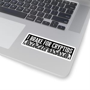 May include: A rectangular sticker with the text "I BRAKE FOR CRYPTIDS" in white on a black background. The sticker features a line of stylized cryptid illustrations in white. The sticker is placed on a laptop.