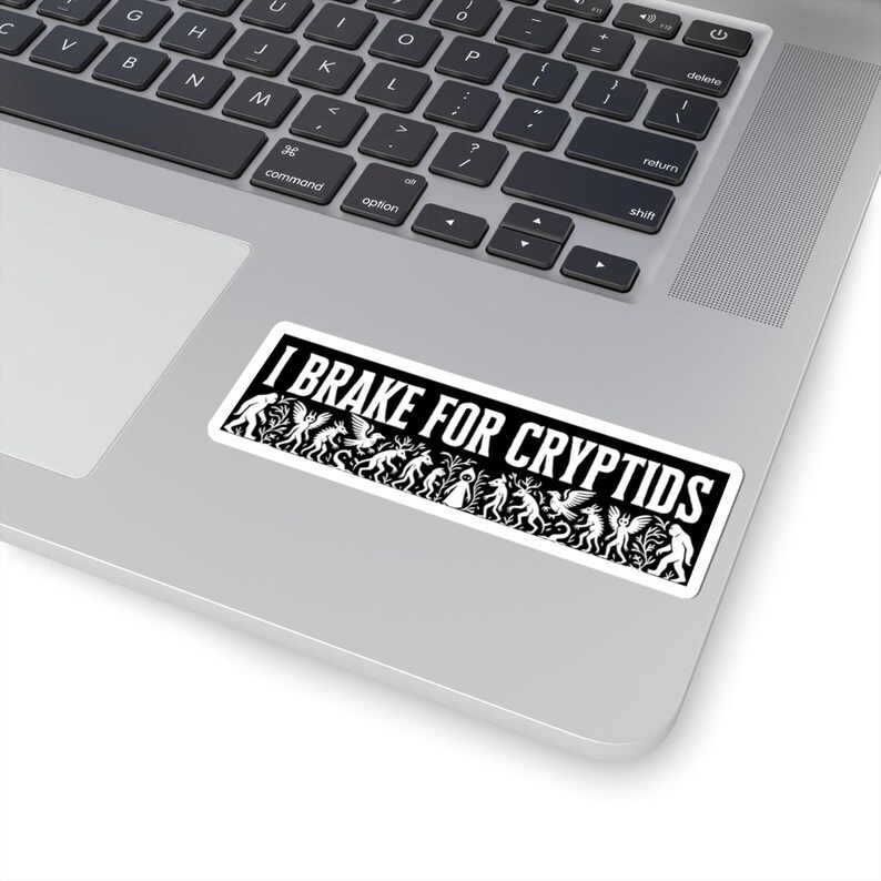 May include: A rectangular sticker with the text "I BRAKE FOR CRYPTIDS" in white on a black background. The sticker features a line of stylized cryptid illustrations in white. The sticker is placed on a laptop.